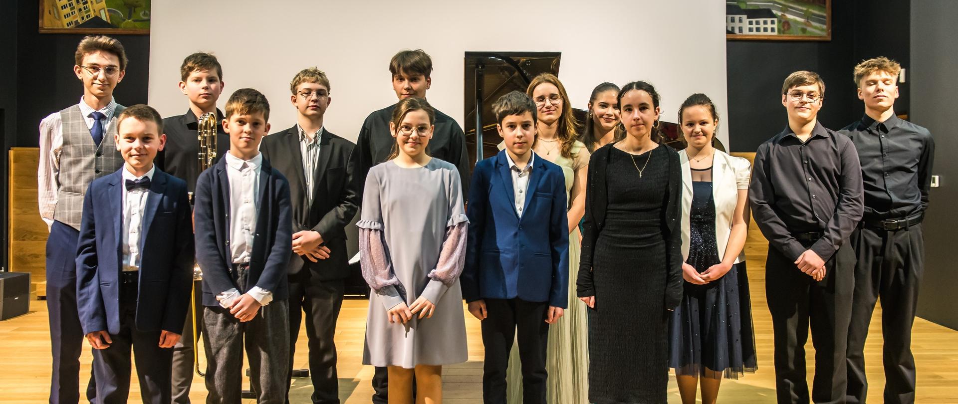 Grupa młodych osób pozuje wspólnie na scenie w eleganckich strojach. W tle widać fortepian oraz fragmenty obrazów zawieszonych na ścianie. Uczestnicy stoją w dwóch rzędach, uśmiechając się do zdjęcia. Jest to pamiątkowe zdjęcie z koncertu szkolnego z dnia 29.01.2026