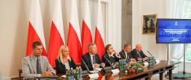 Minister Piontkowski siedzący przy stole w towarzystwie czterech mężczyzn i jednej kobiety. Za nimi pięć flag Polski
