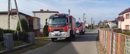 Na zdjęciu znajdują się dwa samochody pożarnicze stojące na drodze na tle domów