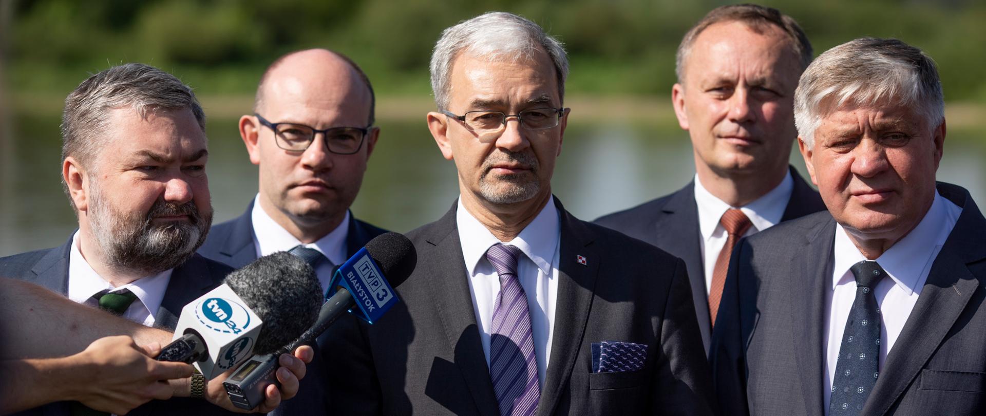 Minister Zieliński stoi przed dziennikarzami, wśród innych uczestników spotkania. W tle widać rzekę. 