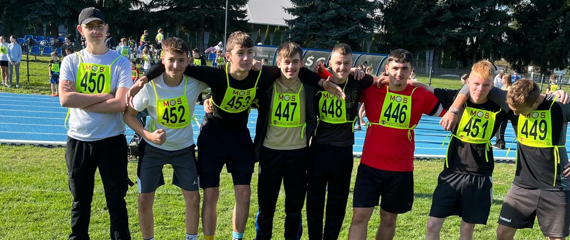 Ośmiu młodych mężczyzn w strojach sportowych i żółtych plastronach startowych stoi w rzędzie na murawie stadionu, obejmując się ramionami.