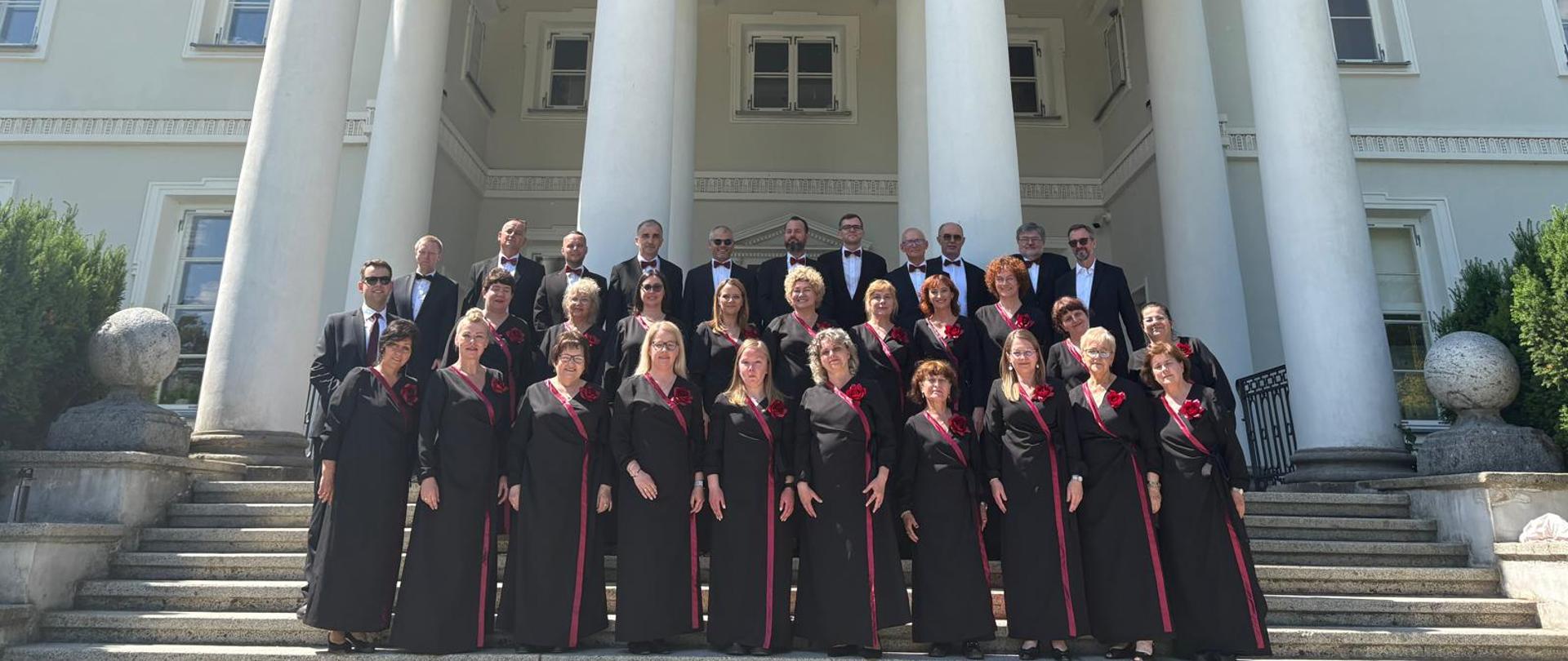 Grupowe zdjęcie chóru wykonane na schodach przed reprezentacyjnym, klasycystycznym pałacem z wysokim portykiem wspartym na białych kolumnach. Członkowie chóru ustawieni są w kilku rzędach – kobiety ubrane są w długie czarne stroje z bordowymi akcentami i ozdobnymi kwiatami, a mężczyźni w ciemne garnitury i muszki. Nad wejściem do budynku widoczny jest łaciński napis umieszczony na fryzie. Fotografia została wykonana w słoneczny dzień przy bezchmurnym niebie i ma uroczysty, pamiątkowy charakter.
