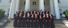 Grupowe zdjęcie chóru wykonane na schodach przed reprezentacyjnym, klasycystycznym pałacem z wysokim portykiem wspartym na białych kolumnach. Członkowie chóru ustawieni są w kilku rzędach – kobiety ubrane są w długie czarne stroje z bordowymi akcentami i ozdobnymi kwiatami, a mężczyźni w ciemne garnitury i muszki. Nad wejściem do budynku widoczny jest łaciński napis umieszczony na fryzie. Fotografia została wykonana w słoneczny dzień przy bezchmurnym niebie i ma uroczysty, pamiątkowy charakter.