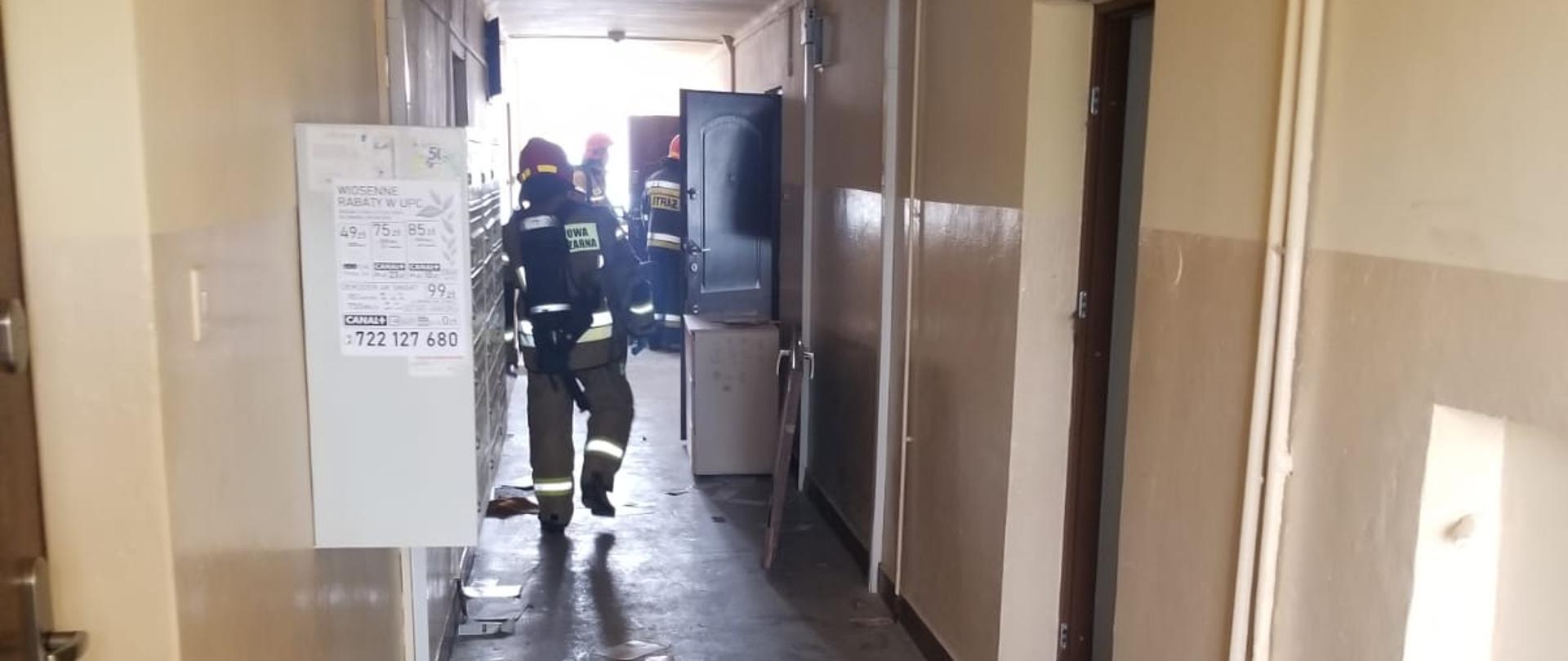 Zdjęcie przedstawia korytarz w budynku mieszkalnym w którym doszło prawdopodobnie do wybuch piecyka gazowego. Na zdjęciu widać strażaków biorących udział w działaniach ratowniczych. Strażacy ubrani są w ubrania specjalne koloru żółtego i granatowego. Na głowach mają czerwone hełmy. Niektórzy z nich dodatkowo posiadają na plechach aparaty ochrony dróg oddechowych. 