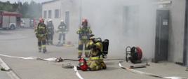 Funkcjonariusze PSP i OSP biorący udział w akcji. Na pierwszym planie strażacy zakładają maski powietrzne przed wejściem do budynku. W tle pojazdy bojowe PSP i OSP oraz wydobywający się dym z pomieszczenia firmy. 