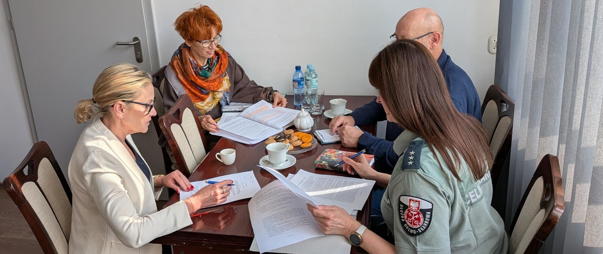 cztery osoby siedzą naprzeciw siebie przy stole, są pochyleni nad dokumentami