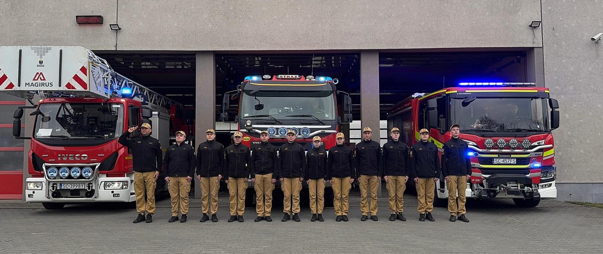 Zastęp strażaków Szkolnej Jednostki Ratowniczo-Gaśniczej stoi przed bramami garażowymi jednostki oddając hołd zmarłemu strażakowi ochotnikowi, w tle stojące samochody bojowe z włączonymi sygnałami świetlnymi