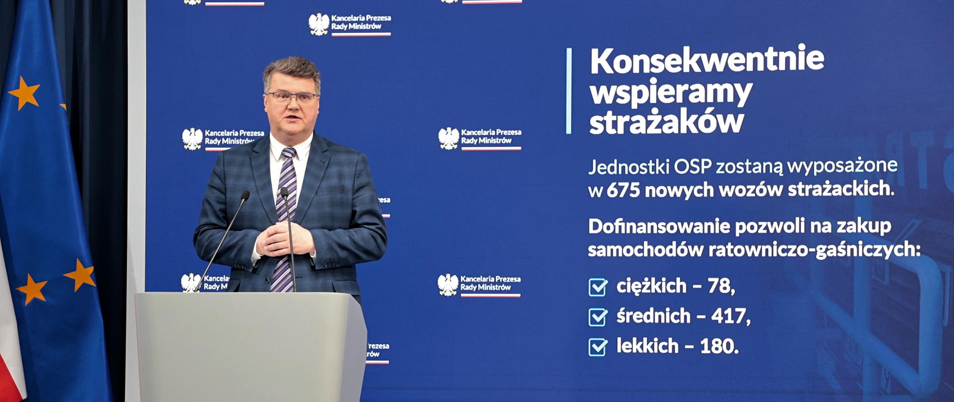 Maciej Wąsik przemawia przy mównicy w trakcie konferencji prasowej za nim banner mswia i napisami konsekwentnie wspieramy strażaków. Jednostki OSP zostaną wyposażone w 675 nowych wozów strażackich. dofinansowanie pozwoli na zakup samochodów ratowniczo-gaśniczych ciężkich 78 średnich 417 lekkich 180