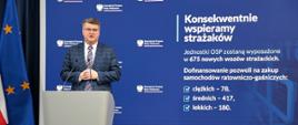 Maciej Wąsik przemawia przy mównicy w trakcie konferencji prasowej za nim banner mswia i napisami konsekwentnie wspieramy strażaków. Jednostki OSP zostaną wyposażone w 675 nowych wozów strażackich. dofinansowanie pozwoli na zakup samochodów ratowniczo-gaśniczych ciężkich 78 średnich 417 lekkich 180