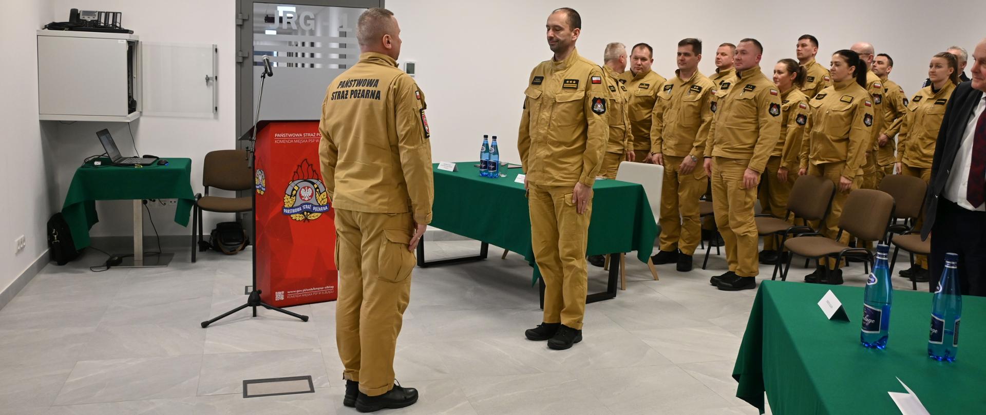 W sali szkoleniowej tutejszej komendy dowódca uroczystości składa meldunek Warmińsko Mazurskiemu komendantowi Wojewódzkiemu. W tle na baczność, stoją strażacy w mundurach