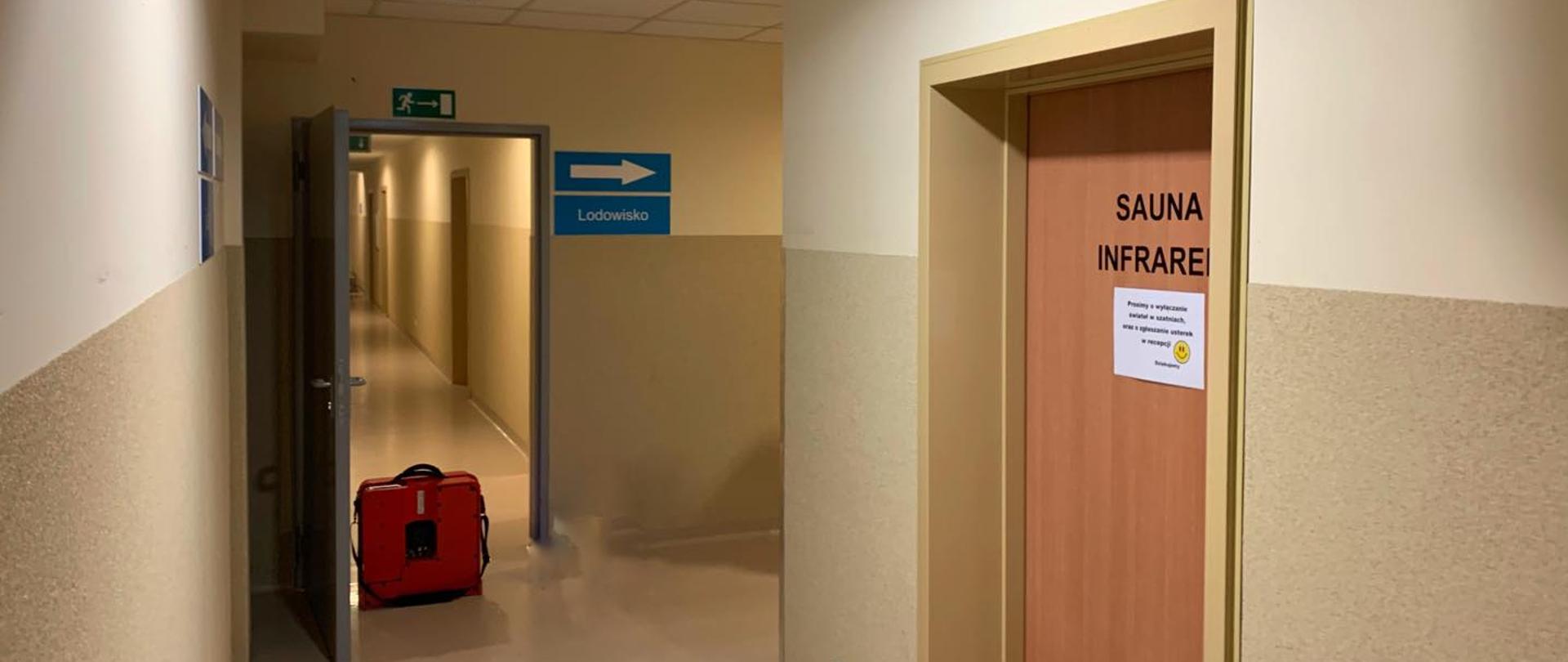 Widok na korytarz. W drzwiach postawiony jest czerwony wentylator oddymiający. Po prawej stronie drzwi a na nich napis SAUNA. 