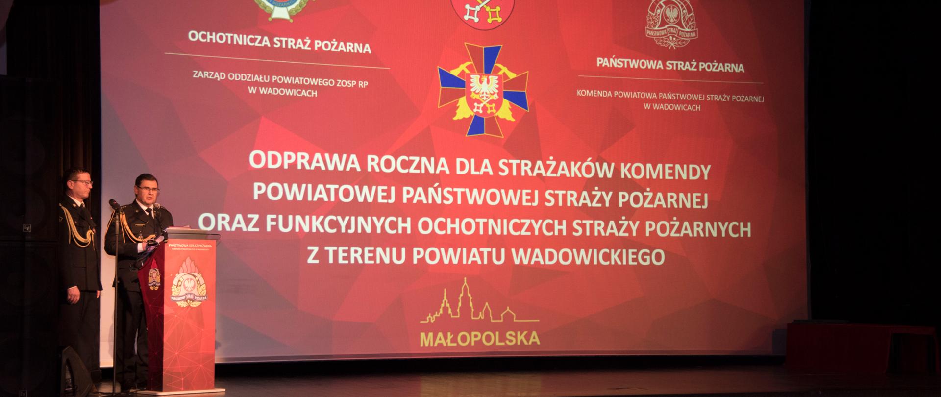 Widok z przodu. Uczestnicy uroczystości siedzą na Sali kinowej WCK, na scenie po lewej stronie Komendant Powiatowy PSP w Wadowicach wita gości, w tle ekran z wyświetloną nazwą wydarzenia.