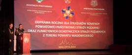 Widok z przodu. Na scenie po lewej stronie Komendant Powiatowy PSP w Wadowicach wita gości, w tle ekran z wyświetloną nazwą wydarzenia.