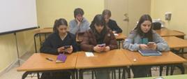 Zdjęcie przedstawia pięcioro młodych uczennic i uczniów w ławkach szkolnych, każdy trzyma w rękach telefon komórkowy jako pomoc naukową. 