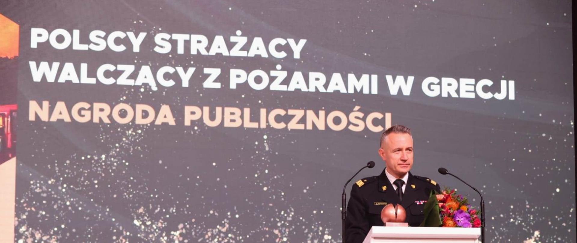 Komendant główny na scenie przy mównicy podczas gali, na mównicy stoi otrzymana statuetka, w tle napis POLSCY STRAŻACY WALCZACY Z POZARAMI W GRECJI NAGRODA PUBLICZNOSCI
