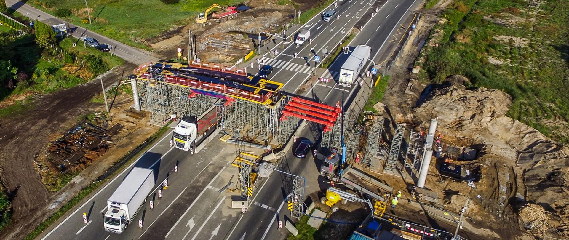 Obecnie priorytetem jest budowa dwóch kładek dla pieszych w Dąbrowie Górniczej, tak aby umożliwić w możliwie najkrótszym czasie bezpieczne przemieszczanie się pieszych nad terenem budowy. Zdjęcie lotnicze obrazuje postęp prac przy budowie jednej z nich. Widoczne na nim liczne filary i podpory obudowane rusztowaniami. Na budowie widoczni pracownicy ubrani z fluorescencyjne ubranie ochronne i wyposażeni w kaski ochronne. Widoczne są również dwie jezdnie istniejące drogi krajowej z wprowadzoną organizacją ruchu na czas robót. 