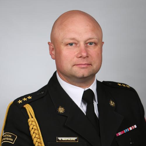 Zdjęcie przedstawia Zastępcę Komendanta Powiatowego PSP w Górze. Jest to portret w ciemnym mundurze strażackim na jasnoszarym tle. Mężczyzna ma ogoloną głowę i patrzy prosto w obiektyw z neutralnym wyrazem twarzy. Na mundurze widoczne są oznaczenia stopnia, odznaczenia oraz tabliczka z nazwiskiem „K. Mastalerz”. Na lewym ramieniu znajduje się ozdobny złoty sznur.”