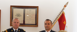 dwóch mężczyzn stoi obok siebie w ciemnych mundurach ze złotym sznurem i białych koszulach z czarnym krawatem. W tle sztandar