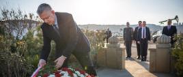 In the photo on the left, Minister Tomasz Siemoniak is adjusting the white-and-red sash on the wreath. The wreath features red (roses), white (gerberas), and green flowers. On the right side of the photo, five men are standing (one in uniform). In the background, you can see oil extraction rigs (derricks), cars, vegetation, and the concrete fence surrounding the memorial site.