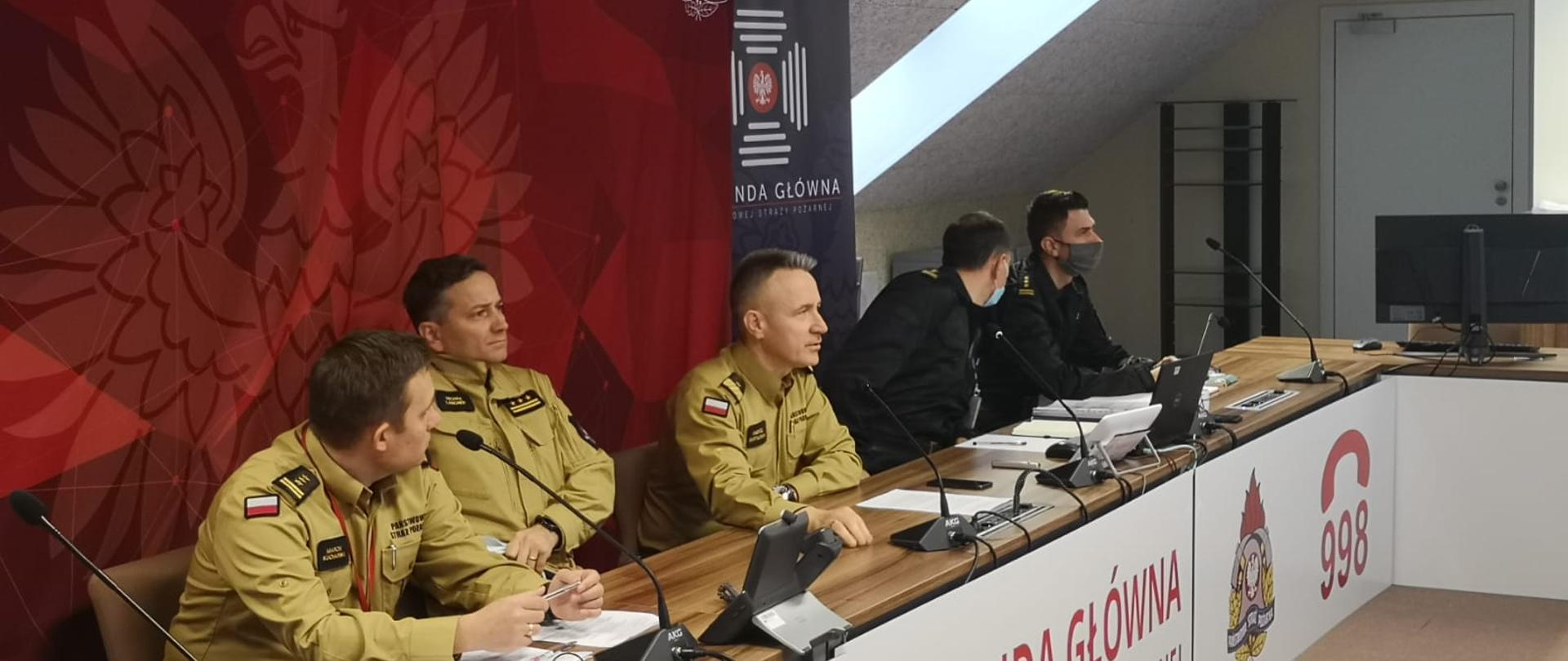 5 strażaków w umundurowaniu , (w środku komendant główny PSP) siedzą przy biurku podczas wideokonferencji, za nimi ustawione ścianki z wizerunkiem godła Polski oraz logotypu KG PSP