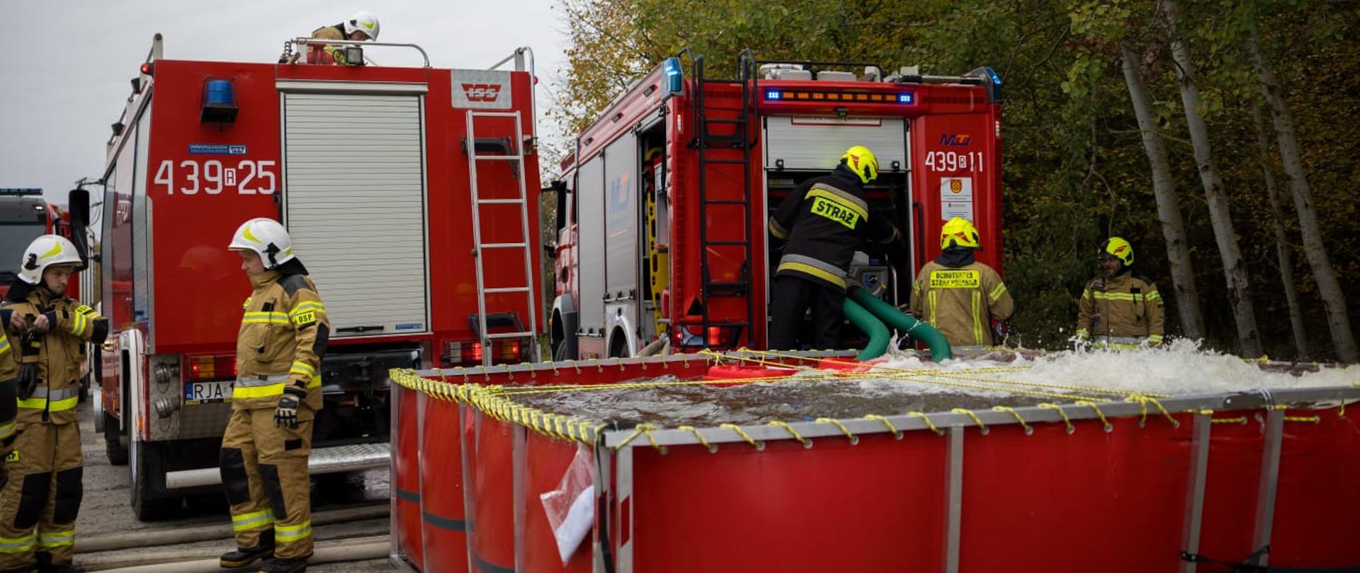 Strażacy podczas ćwiczeń z wykorzystaniem samochodów pożarniczych i zbiornika na wodę.