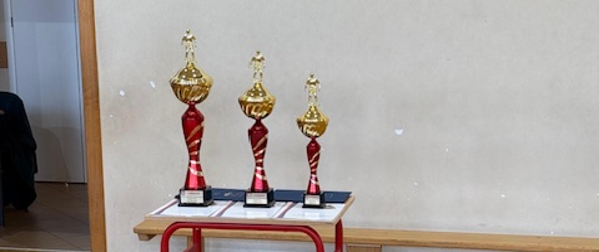 Trzy puchary na czerwonym stoliku, stojące na drewnianej podłodze w pomieszczeniu. Puchary są w różnych rozmiarach, prawdopodobnie symbolizując pierwsze, drugie i trzecie miejsce. Każdy puchar ma czerwoną podstawę i złotą górę. Tło stanowi jasna ściana z czarnym paskiem u dołu. Po lewej stronie widoczne są drzwi. Na drewnianej podłodze widoczne są linie.