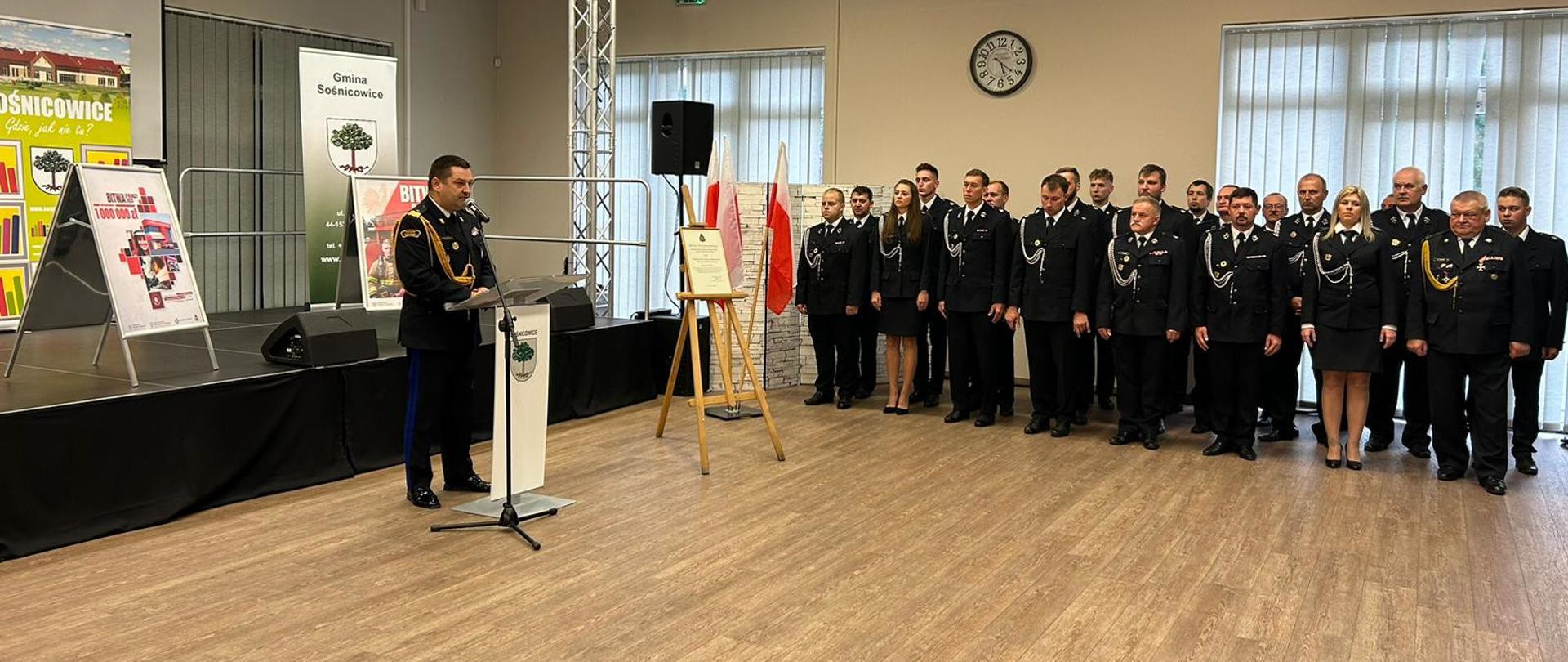 Sala konferencyjna, przed sceną na której ustawione są banery gminy Sośnicowice oraz akcji "Bitwa o remizy" stoją w za mównicą przemawia śląski komendant wojewódzki PSP nadbryg. Jacek Kleszczewski, po prawej stronie w dwóch rzedach stoją w mundurach wyjściowych druhowie z ochotniczych straży pożarnych.