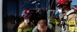 Fotografia przedstawia działania strażaków związane z ewakuacją osoby poszkodowanej z uszkodzonego pojazdu. W centralnej części kadru ratownicy prowadzą poszkodowanego przez otwór w pojeździe, wykorzystując odpowiednie zabezpieczenie osobiste oraz sprzęt przeznaczony do działań ratowniczych.
Widoczne uszkodzenia pojazdu, w tym rozbita szyba, wskazują na charakter realizowanego scenariusza, obejmującego zdarzenie komunikacyjne. Ratownicy zabezpieczają poszkodowanego podczas opuszczania pojazdu, jednocześnie zachowując kontrolę nad jego przemieszczaniem. Zdjęcie dobrze obrazuje współpracę członków zastępu podczas prowadzenia ewakuacji z przestrzeni o ograniczonym dostępie.