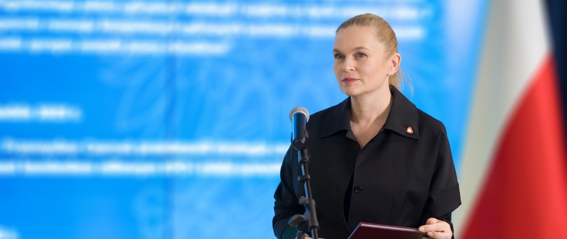 Minister Nowacka stoi i przy mikrofonie w tle duży ekran z prezentacją
