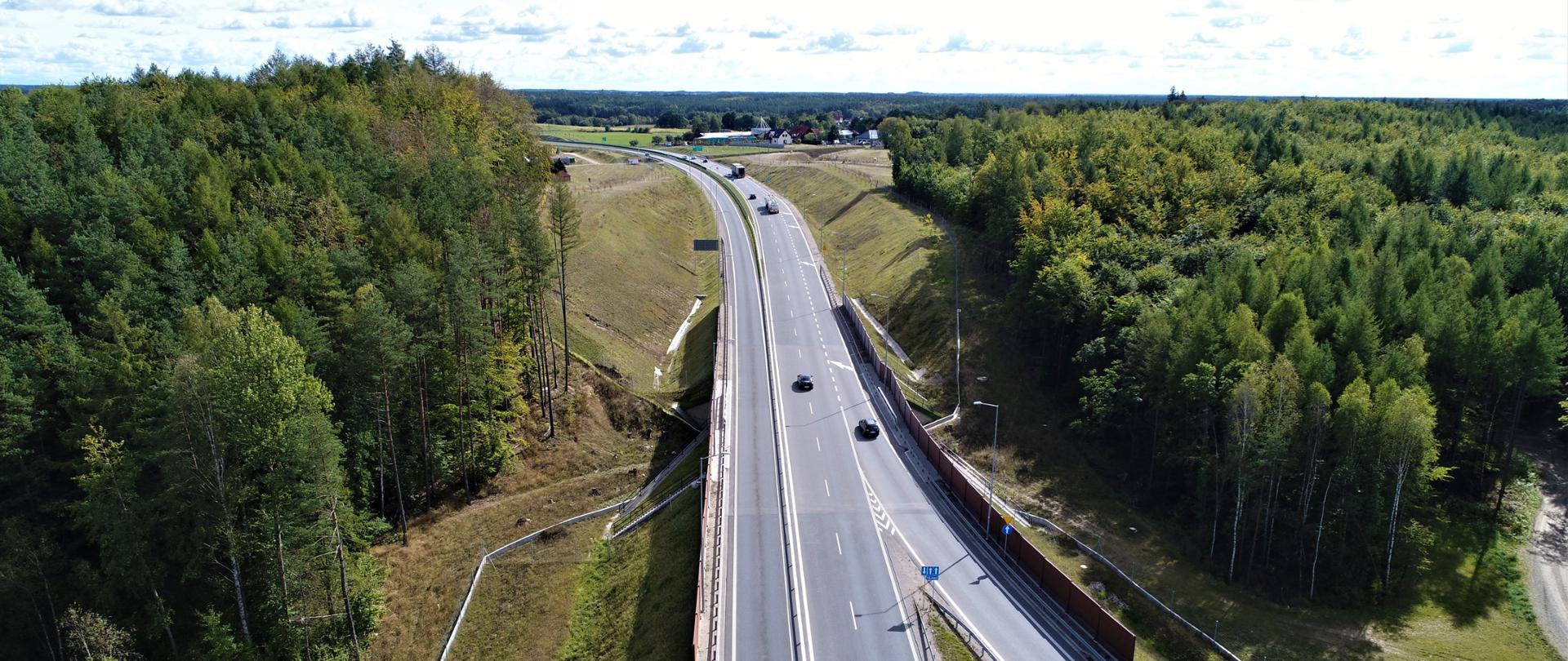 Zdjęcie ilustracyjne obwodnicy. W centrum zdjęcia droga z pojazdami, po bokach fragmenty obszar porośniętego drzewami.