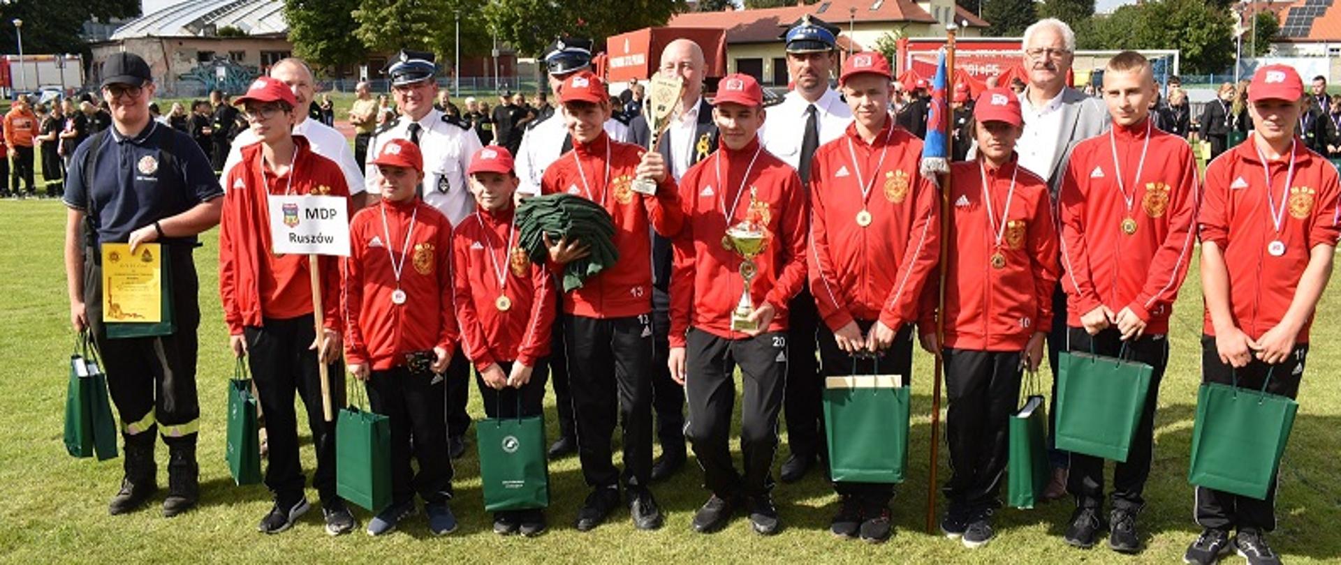 Dzień Na murawie boiska stoi grupa dzieci ubrana w czarne spodnie dresowe, czerwone bluzy i czerwone czapki z daszkiem. Jedno z dzieci trzyma tabliczkę z napisem MDP Ruszów. Dzieci trzymają w rękach zielone papierowe torby. Dwoje z dzieci ma w rękach dwa puchary. Obok dzieci stoi mężczyzna ubrany w czarne spodnie od munduru koszarowego, granatową koszulę typu "polo" i czarną czapkę z daszkiem. W ręce trzyma zieloną torbę oraz dyplom w kolorze żółtym. Za dziećmi stoi kilku funkcjonariuszy PSP w białych koszulach i rogatywkach oraz kilka osób cywilnych. W tle zdjęcia widać ustawione w szeregu dzieci.
