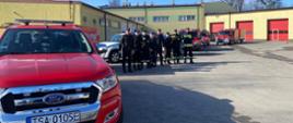 Zdjęcie zrobione na placu. Widać strażackie samochody z przyczepkami i ośmiu strażaków stojących obok nich.