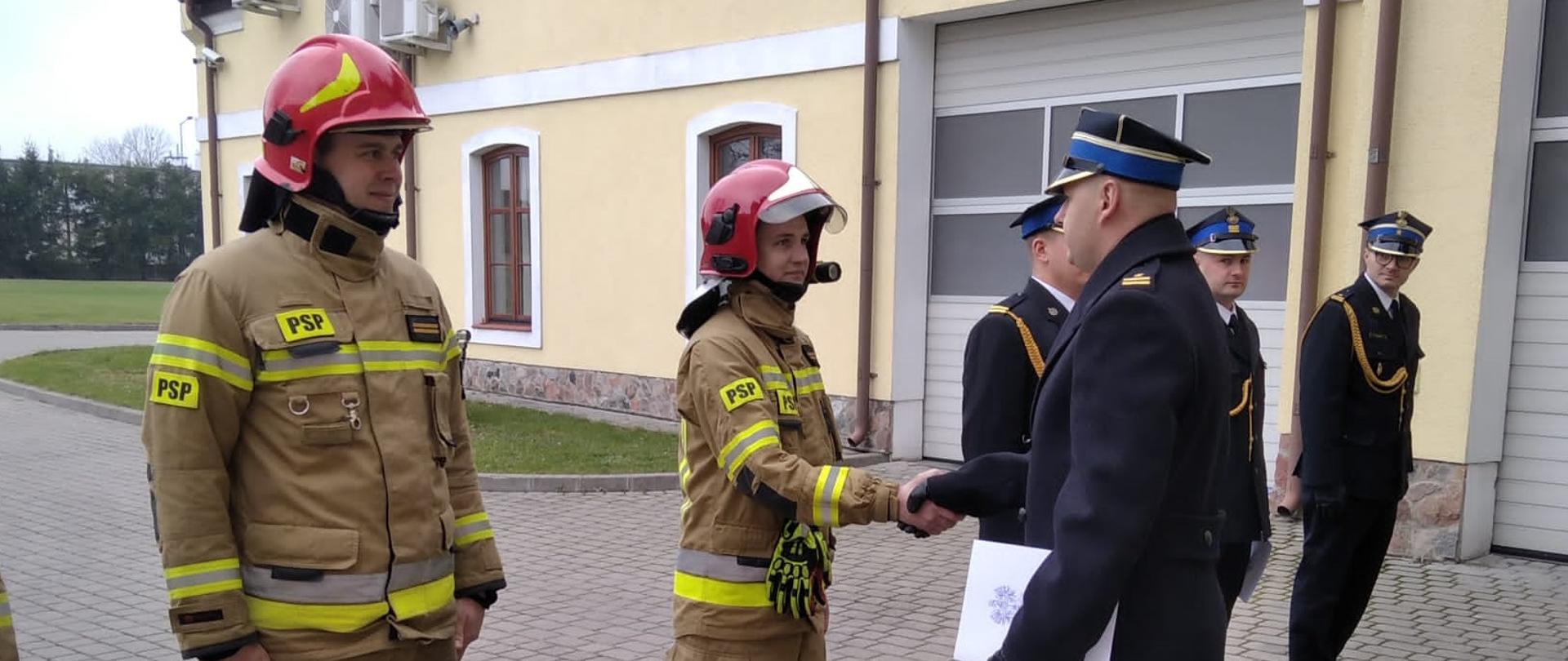 Strażacy stoją na uroczystej zbiórce. Komendant wręcza im awanse. 2 strażaków ubranych jest w ubranie specjale a 3 w mundur. 