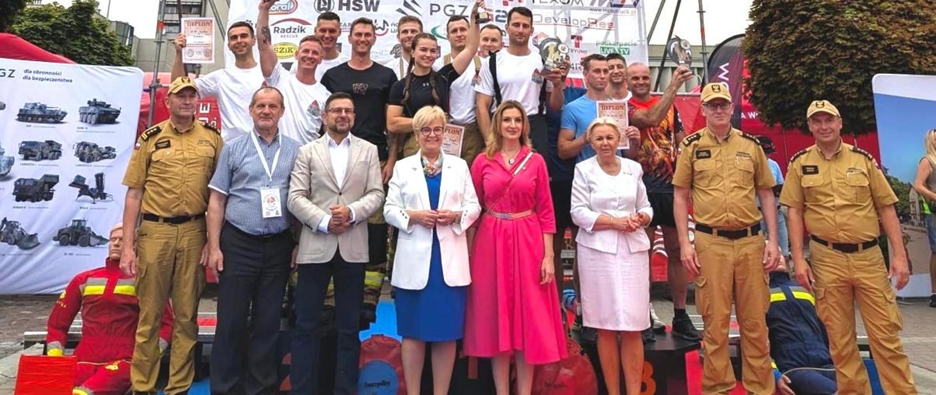 Zdjęcie grupowe przedstawiającego strażaków reprezentujących KP PSP w Jarosławiu wraz ze swoimi przełożonymi oraz kierownictwem zawodów . Strażacy ubrani w pamiątkowe koszulki. Zdjęcie wykonane na placu zawodów. W tle widoczny tor przeszkód.