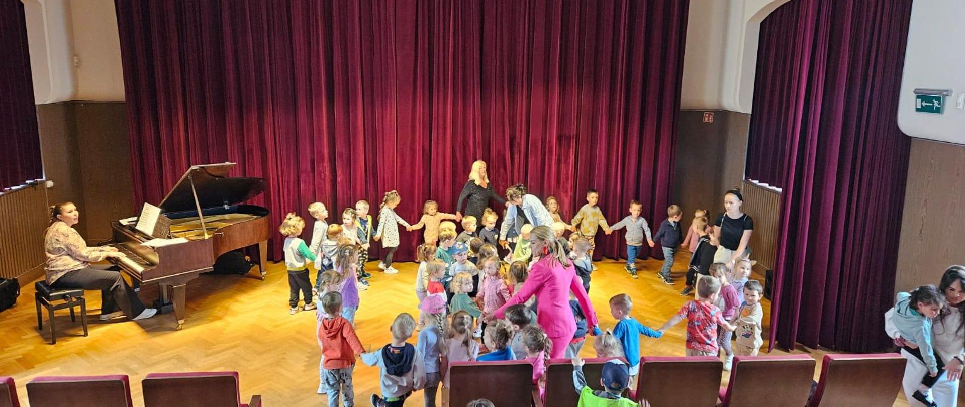 Na środku grupa bawiących się dzieci, a po lewej stronie na fortepianie gra p. Iryna Yakymets