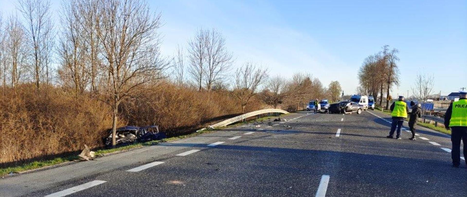 Zdjęcie przedstawia szeroką perspektywę miejsca gdzie doszło do zderzenia dwóch samochodów osobowych. Po lewej stronie, w rowie leży jeden ze zniszczonych aut. Drugi z pojazdów stoi na środku drogi. W tle radiowozy policyjne. Na drodze stoją funkcjonariusze Policji.
