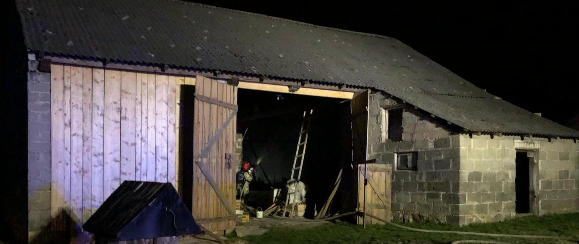 Zdjęcie przedstawia stodołę, w której doszło do pożaru: przez otwarte wrota widać strażaka podającego wodę na drewnianą konstrukcję budynku.