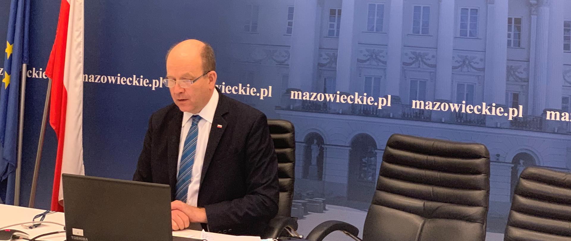 Wojewoda Mazowiecki Konstanty Radziwiłł prowadzący posiedzenie online zespołu zarządzania kryzysowego. Siedzi przy stole, na którym znajduje się laptop. W tle granatowa ścianka ze zdjęciem siedziby Mazowieckiego Urzędu Wojewódzkiego.