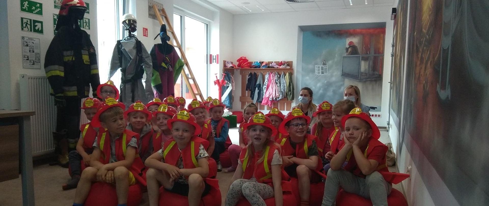 Dzieci siedzą na pufkach w sali ognik. Są ubrane w kamizelki odblaskowe oraz kaski strażackie.