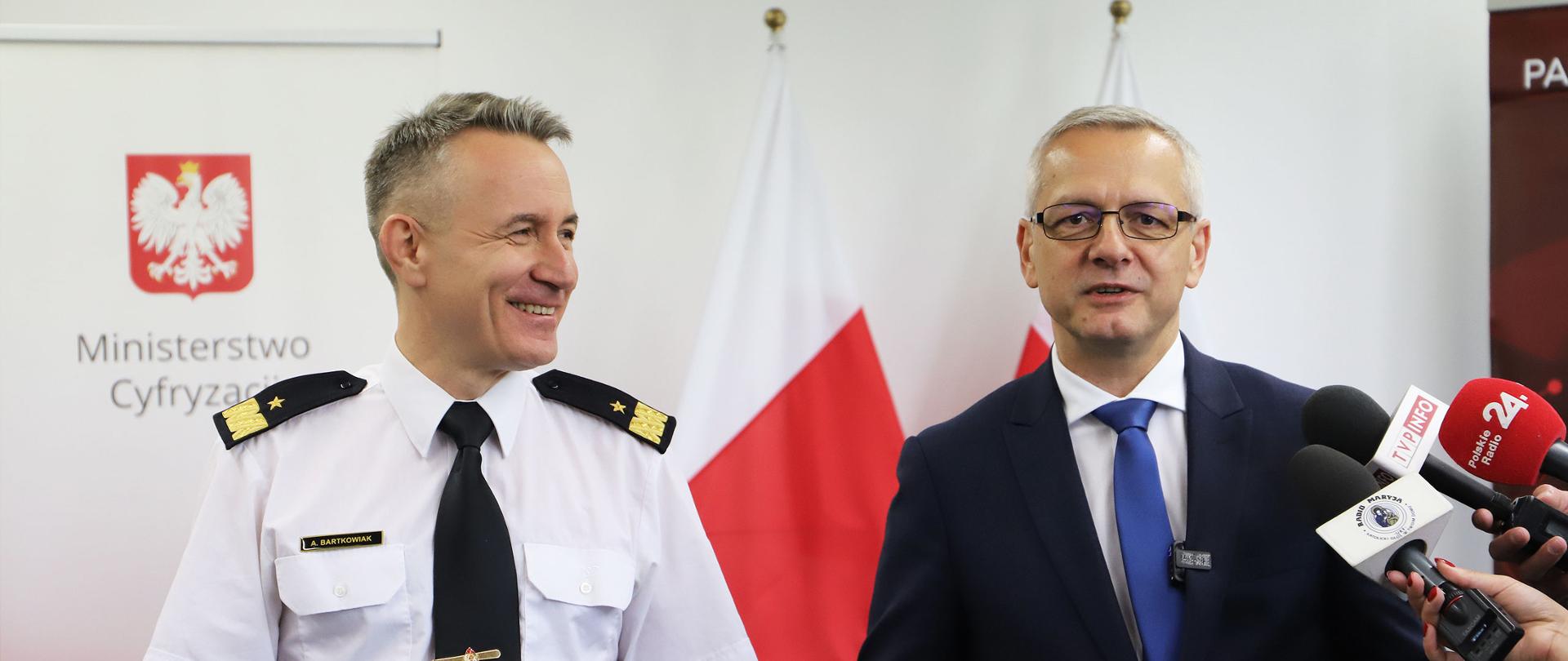 Minister cyfryzacji i komendant główny Państwowej Straży Pożarnej w trakcie briefingu prasowego. W tle roll - up z napisem Ministerstwo Cyfryzacji i polskie flagi.