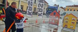 Strażak wraz z dzieckiem w hełmie strażackim podają wodę z węża na makietę domku. W tle tarnowskie kamienice oraz flagi promujące jazdę w kaskach.