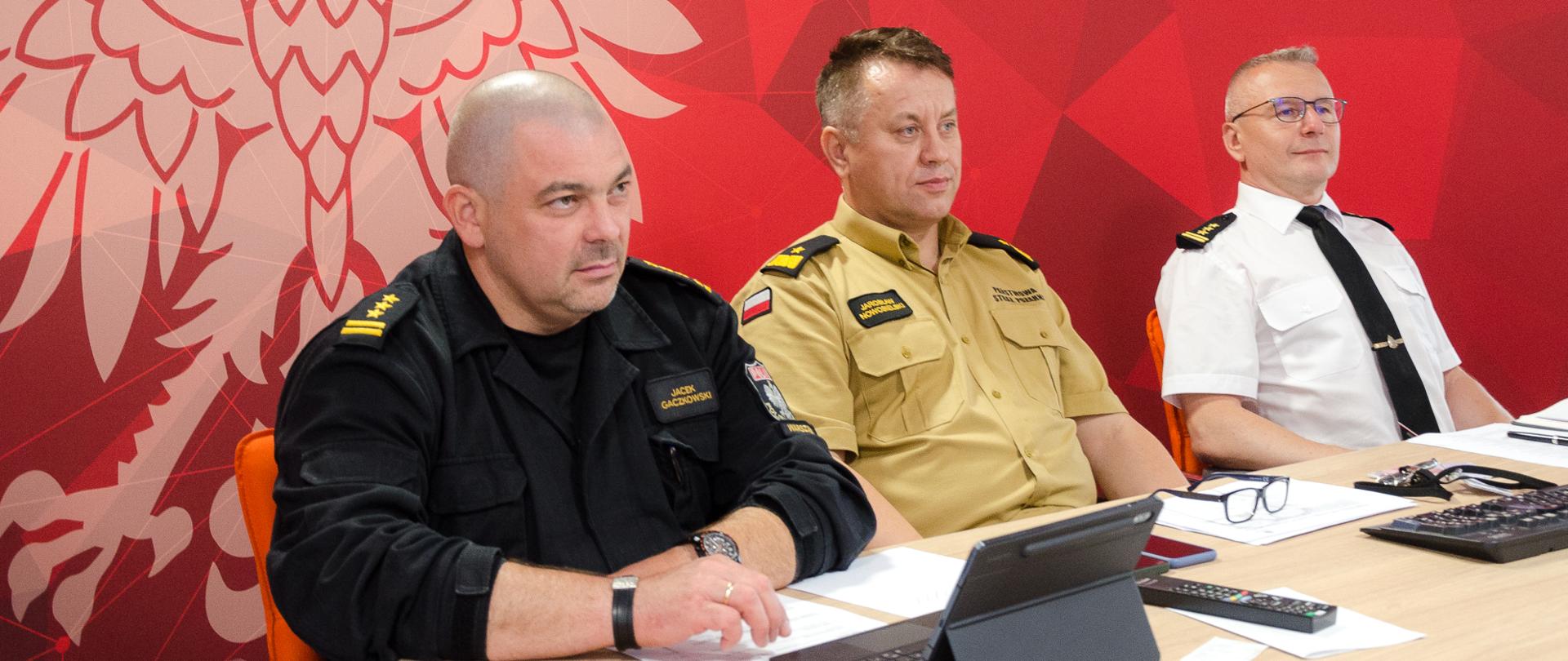 Na zdjęciu widać trzech strażaków podczas wideokonferencji. Siedzą za stołem, w tle banner koloru czerwonego.