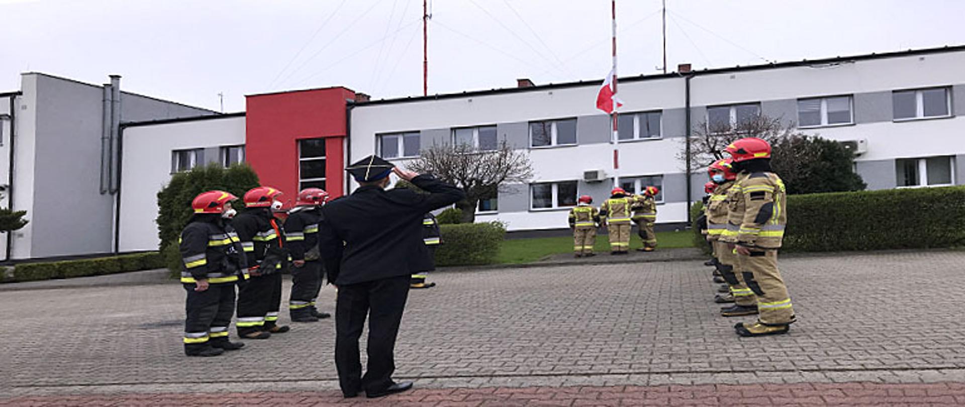 Zdjęcie przedstawia zmianę służbową strażaków podczas podnoszenia flagi państwowej na maszt. Na czele szeregu na pierwszym planie stoi oficer w mundurze wyjściowym z czapką rogatywką i salutuje. Strażacy stoją frontem do siebie w szeregu dziewięciu na lewej stronie i ośmiu po prawej stronie, wszyscy spoglądają na maszt, na który jest wciągana flaga państwowa przez trzech ratowników. Jeden strażak salutuje podnoszonej na maszcie fladze, po jego lewej stronie strażak stoi w pozycji zasadniczej, po prawo ratownik wciąga flagę, flaga znajduje się w połowie długości masztu. Wszyscy strażacy są ubrani w ubrania bojowe typu Moratex, na głowie maja heł, na twarzy maseczki ochronne, na nogach kalosze. Ratownicy po lewo ubrani są w ubrania barwy czarnej z odblaskowymi pasami barwy szarej i żółtej a ratownicy po prawej stronie kadru w ubrania barwy jasno brązowej z odblaskowymi pasami barwy szarej i żółtej. W tle żywopłot oraz budynek Komendy Powiatowej w Łowiczu oraz okna należące do części administracyjnej komendy.