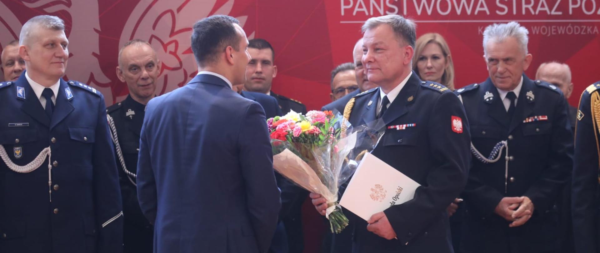 Na zdjęciu Dyrektor Biura Wojewody Jarosław Draguć wręcza w imieniu Wojewody Opolskiego list gratulacyjny st. bryg. Pawłowi Kielarowi, nowemu Opolskiemu Komendantowi Wojewódzkiej Państwowej Straży Pożarnej w Opolu. St. bryg. Paweł Kielar trzyma w ręku kwiaty oraz otrzymany list gratulacyjny. W tle widać pozostałych uczestników uroczystości.