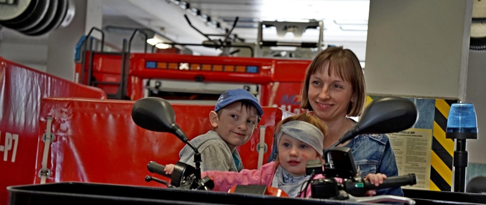 Na zdjęciu widoczna mam z dwójką dzieci dziewczynką i chłopcem siedzą we trójkę na strażackim czerwonym quadzie który znajduje się na garażu w Komendzie Powiatowej Państwowej Straży Pożarnej w Gorlicach