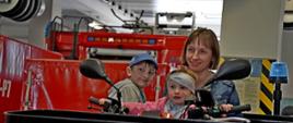 Na zdjęciu widoczna mam z dwójką dzieci dziewczynką i chłopcem siedzą we trójkę na strażackim czerwonym quadzie który znajduje się na garażu w Komendzie Powiatowej Państwowej Straży Pożarnej w Gorlicach