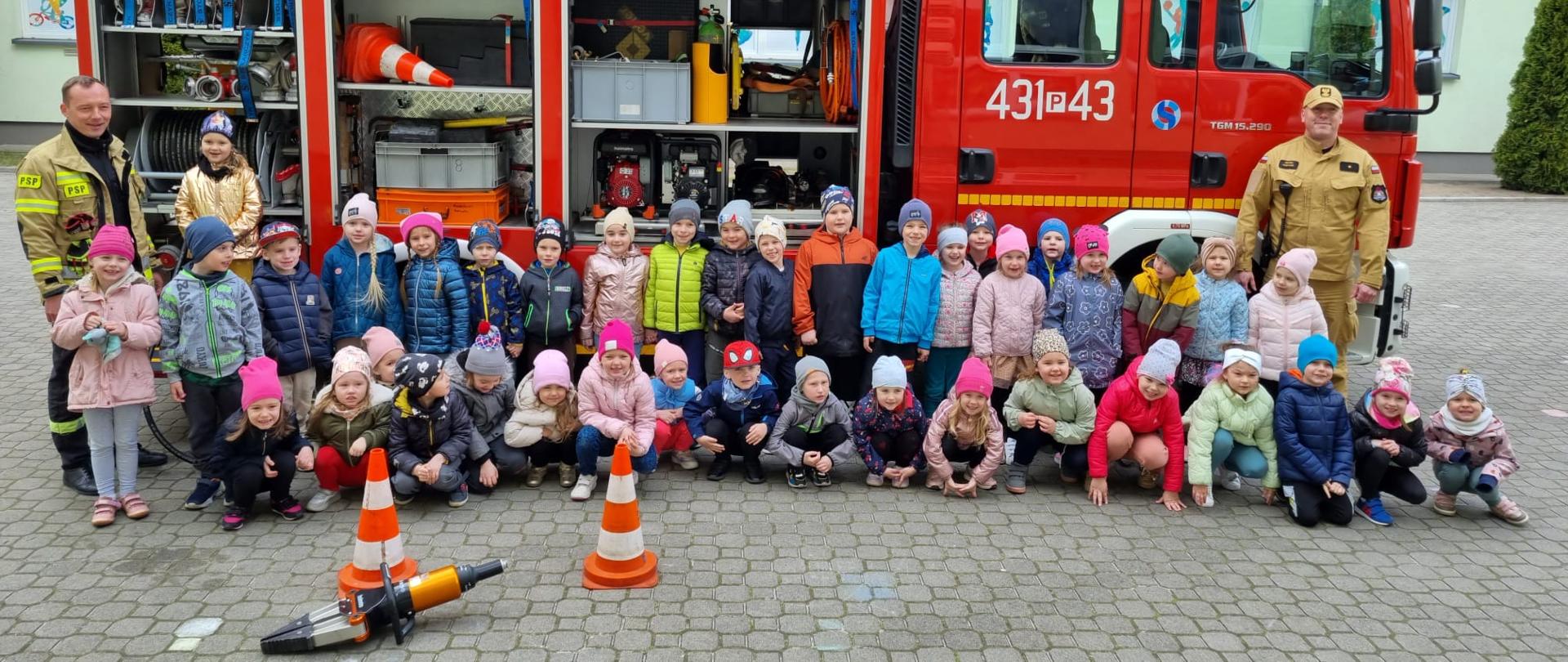 Zdjęcie przedstawia dwóch strażaków oraz grupę dzieci, którzy pozują do zdjęcia na tle wozu strażackiego. Skrytki samochodu są otwarte, przed grupą dzieci leży rozpieracz hydrauliczny.