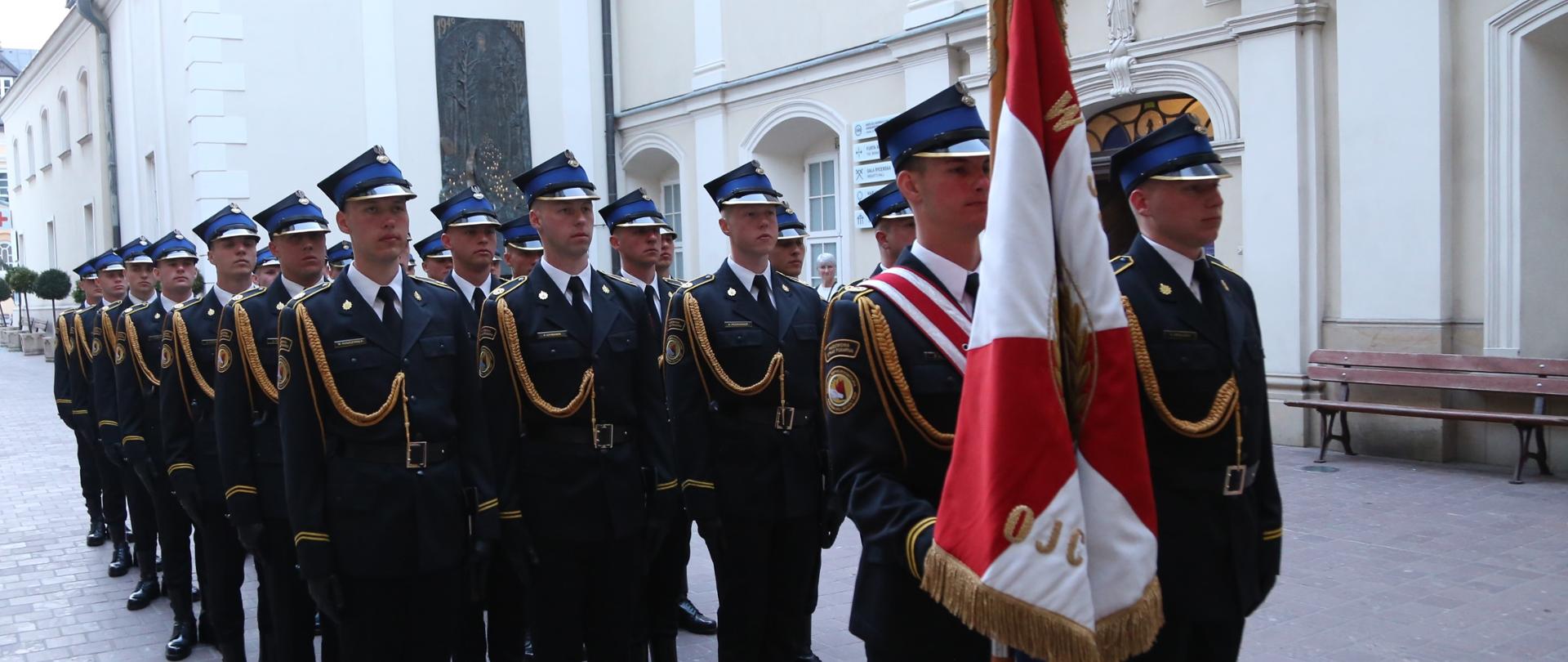 Stojąca w czterech rzędach kompania honorowa Centralnej Szkoły PSP na czele której stoi poczet sztandarowy złożony z dwóch kadetów CS PSP z których jeden w mundurze galowym przepasany biało-czerwoną wstęgą trzyma sztandar