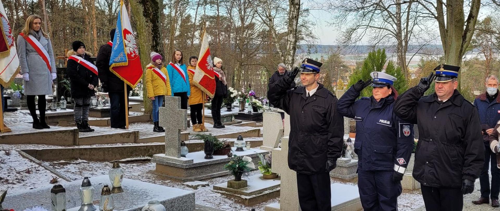 Zdjęcie przedstawia poczty sztandarowe podczas uroczystego capstrzyka przy powstańczej mogile.
W tle drzewa i groby na cmentarzu.
