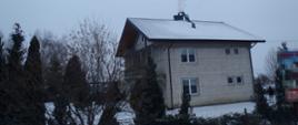Zdjęcie przedstawia budynek mieszkalny – dom jednorodzinny wykonany z białej cegły pokryty blachą. Na dachu przy kominie znajduje się jeden strażak który dokonuje czyszczenia przewodu kominowego.